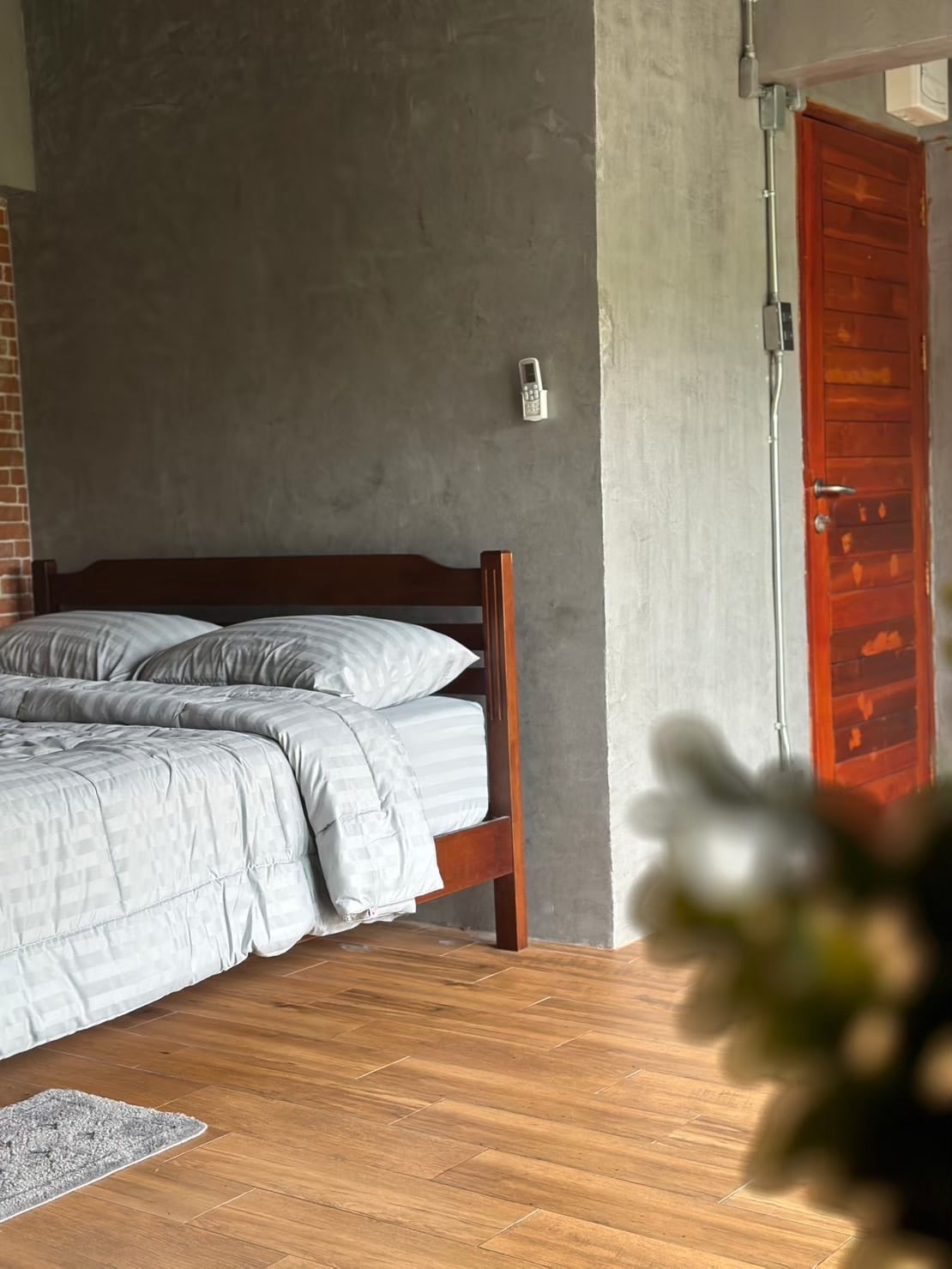 A simple and well-maintained bedroom features a wooden bed with light grey bedding. The room showcases a textured wall in neutral tones and has a wooden door that adds warmth to the space. Natural light brightens the area, enhancing a comfortable ambiance.