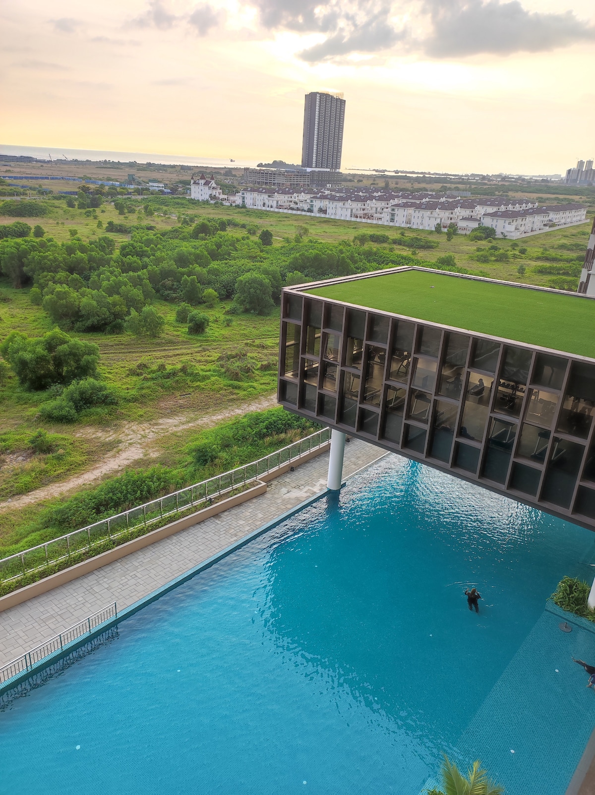 A spacious outdoor area features a large swimming pool with clear blue water and a green rooftop terrace. The surrounding landscape includes greenery and residential buildings in the distance, offering a serene view.