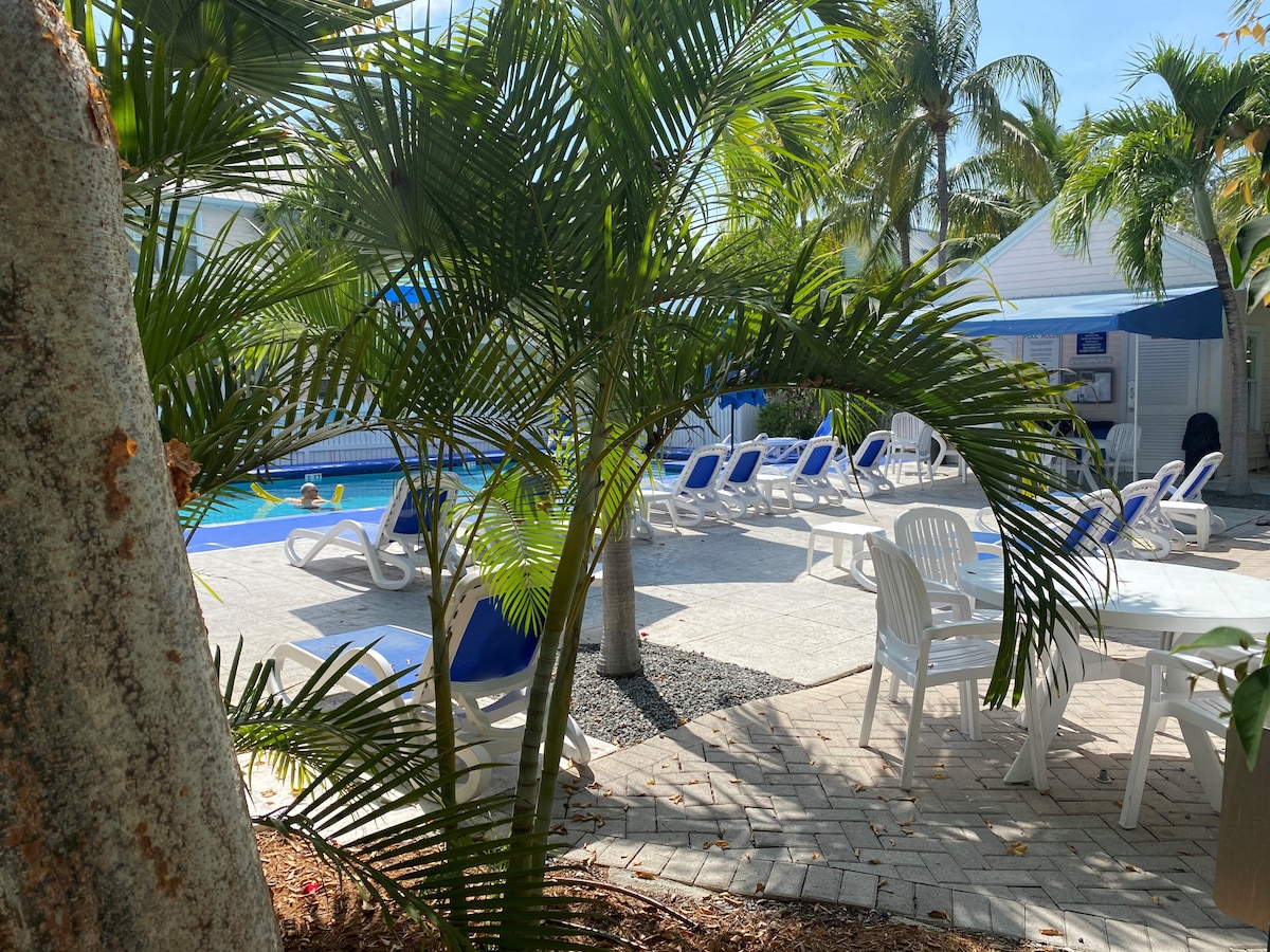 A community pool is surrounded by palm trees and lounge chairs. The pool area is paved with light-colored tiles and features a shaded seating area. Several guests can be seen enjoying the water under the bright sky.