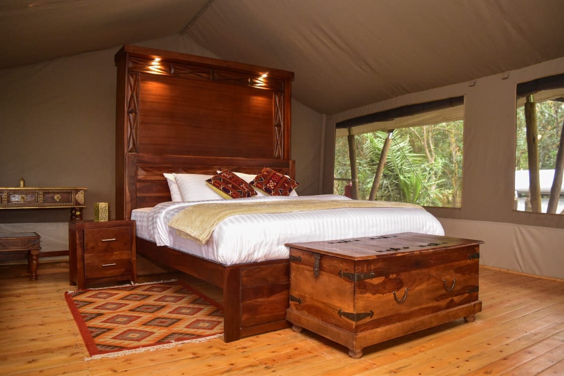 A spacious sleeping area features a wooden bed with an elaborately carved headboard, complemented by quality bedding and decorative pillows. A rustic chest sits at the foot of the bed. Natural light floods in through large windows framed by green surroundings.