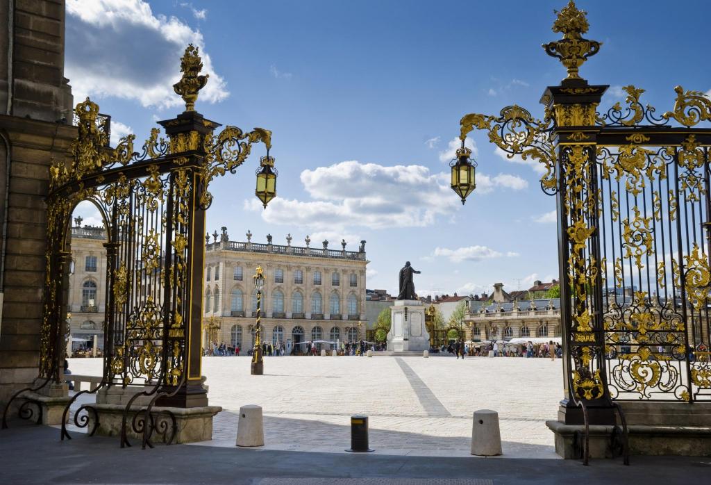 The image captures the view through ornate golden gates, leading to a spacious square. In the background, a large building with multiple windows and a statue in the center is visible. Bright blue skies and scattered clouds enhance the overall scene.