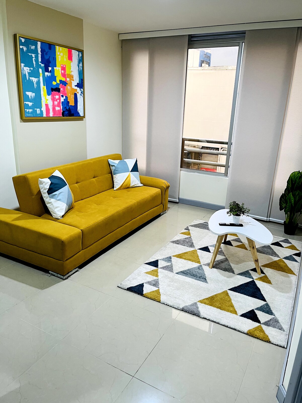 A bright living area is presented, featuring a yellow sofa adorned with decorative pillows. A small round coffee table sits on a geometric patterned rug. Natural light fills the space through large windows covered by blinds, creating a welcoming environment.