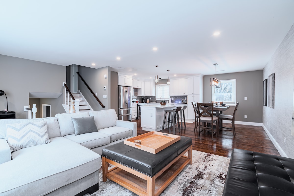 A spacious living area combines a cozy sectional sofa and a sleek coffee table with a light wood finish. Adjacent, a dining table accommodates six, and a modern kitchen features stainless steel appliances. Soft lighting enhances the welcoming atmosphere of the open layout.