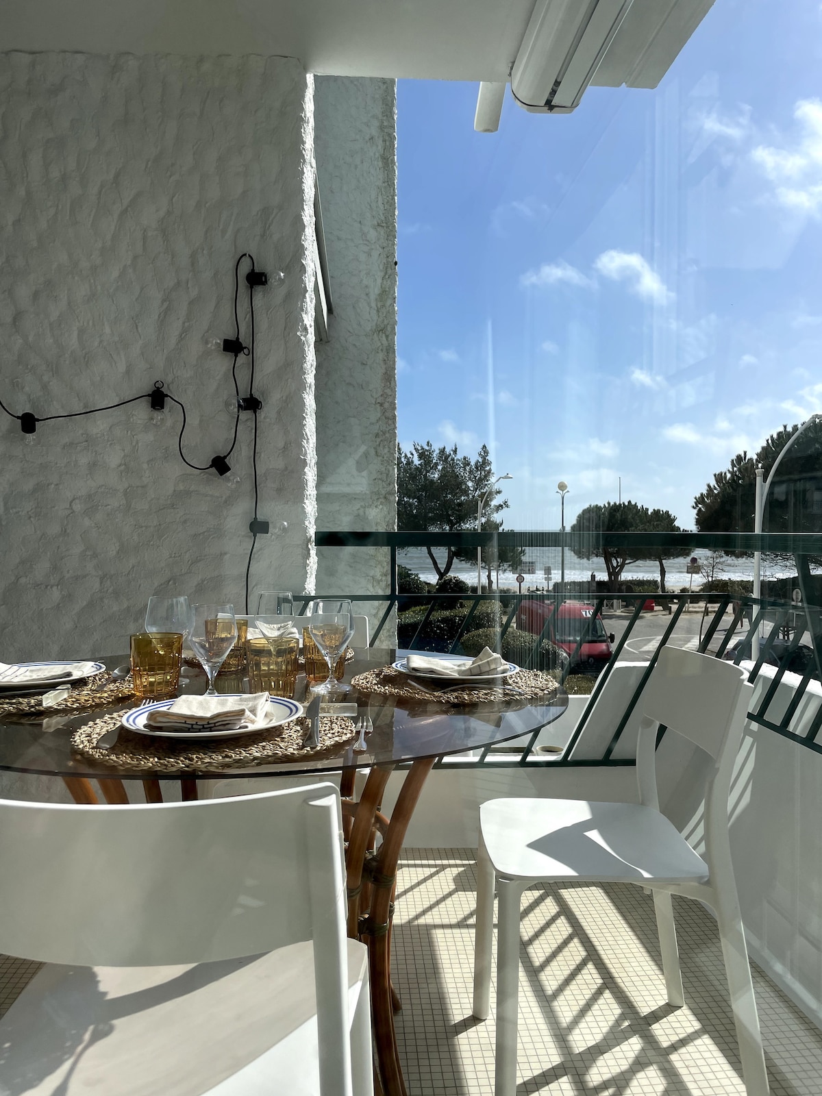 A dining area on the terrace features a round table set for six, adorned with glassware and woven placemats. The backdrop highlights a clear sky and coastal views, with sunlight filtering through large glass doors, creating a bright and inviting space.