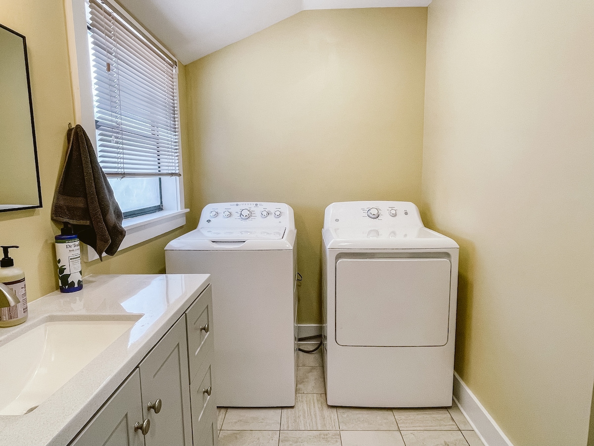 A laundry room is equipped with a washer and dryer, positioned side by side against a light yellow wall. Natural light enters from a window with blinds, illuminating a clean countertop below the mirror. Gray cabinetry offers storage space, complementing the light-colored tiled floor.