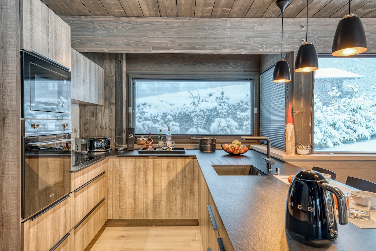 A modern kitchen features sleek wooden cabinetry and a black countertop. A large window provides a view of the snowy outdoors. Kitchen appliances, including an electric kettle and toaster, are arranged neatly on the counter, contributing to a spacious and functional design.
