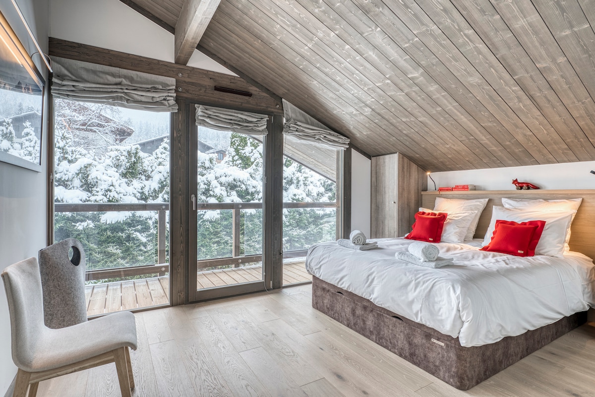 A serene bedroom features a spacious bed with white linens and red accent pillows. Large windows provide views of the snowy landscape outside, allowing natural light to fill the room. A simple chair is positioned nearby on the light-colored wooden floor.
