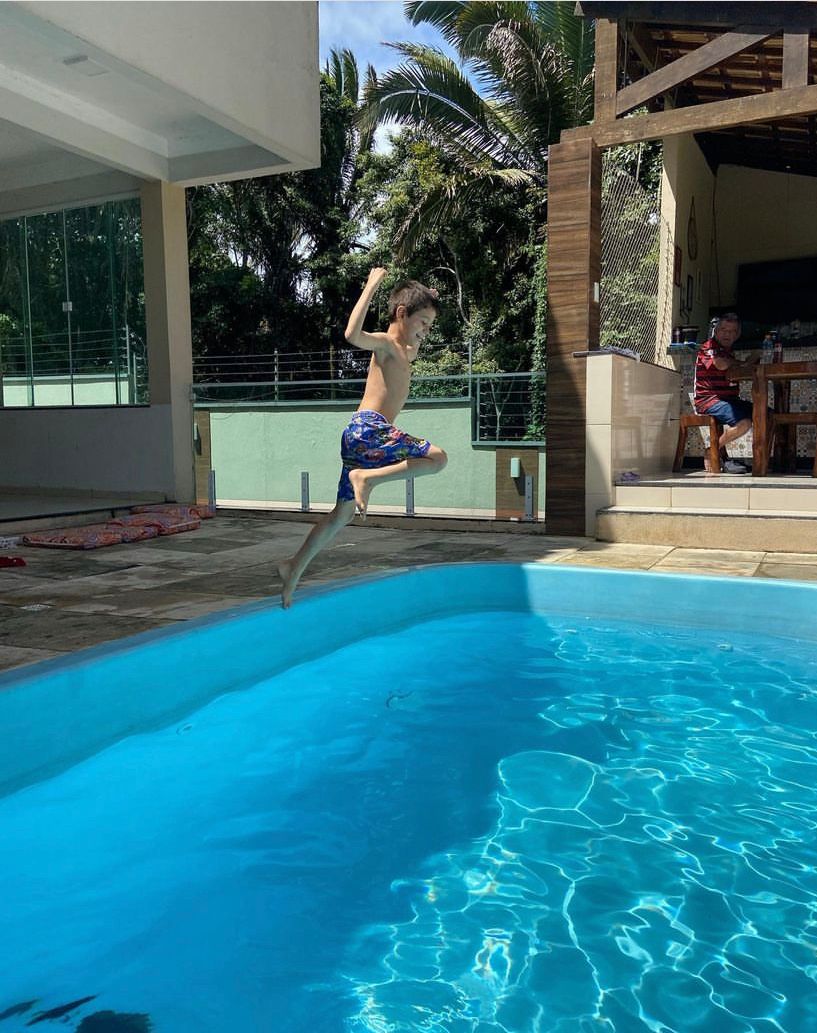 A child is captured mid-air, joyfully leaping into a clear blue swimming pool. Surrounding the pool, a spacious patio area is visible, with tropical foliage and a dining set partially seen in the background.