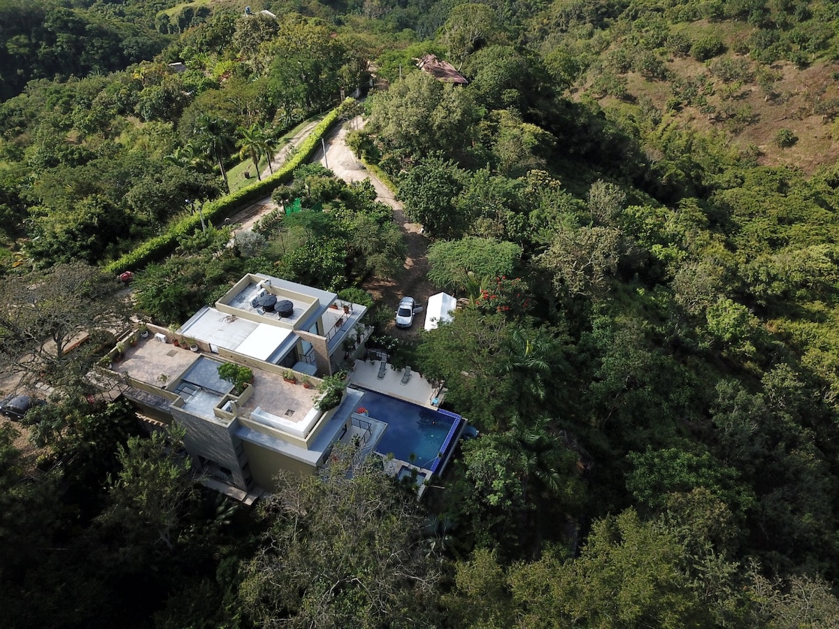 An aerial view presents a modern rural house surrounded by lush greenery, highlighting a spacious terrace with a pool. Nearby, a winding unpaved road is visible, leading into the dense landscape. The exterior design complements the natural surroundings.