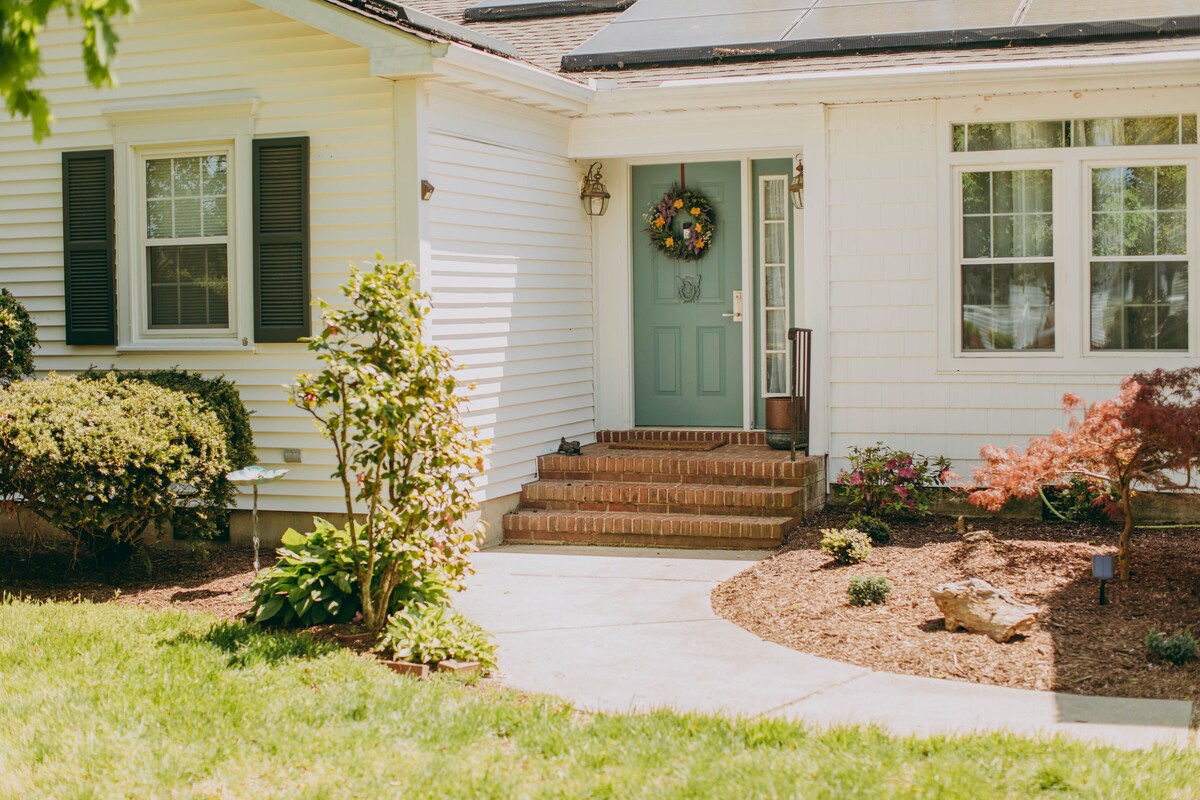 The entrance to the home features a welcoming front door adorned with a wreath. Lush greenery and landscaped garden beds surround the path, which leads to a set of brick steps. A mixture of plants, including a small tree, enhances the front yard.