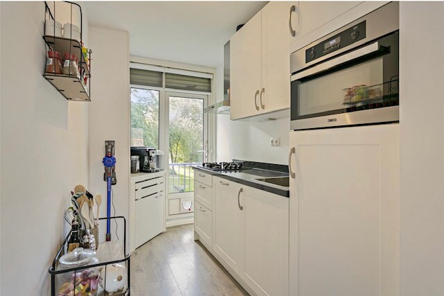 A modern kitchen features sleek white cabinetry and a dark countertop. Natural light enters through the large window, illuminating the prep area. Small appliances, including a coffee maker, are arranged on the countertop, while shelf storage displays kitchen essentials.
