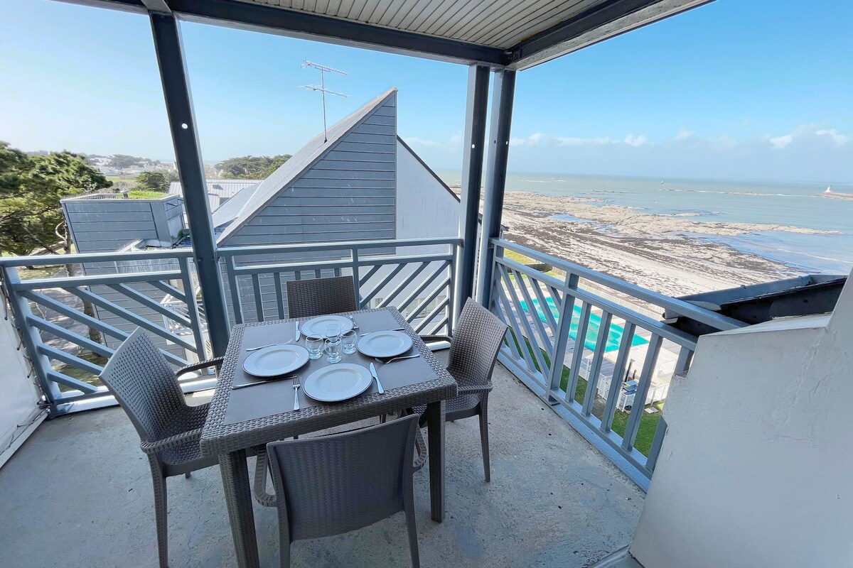 A terrace is presented with a dining table set for four, offering ocean views. Surrounding greenery and the coastline can be seen in the background. The space features a shaded overhang, providing a comfortable outdoor dining area.