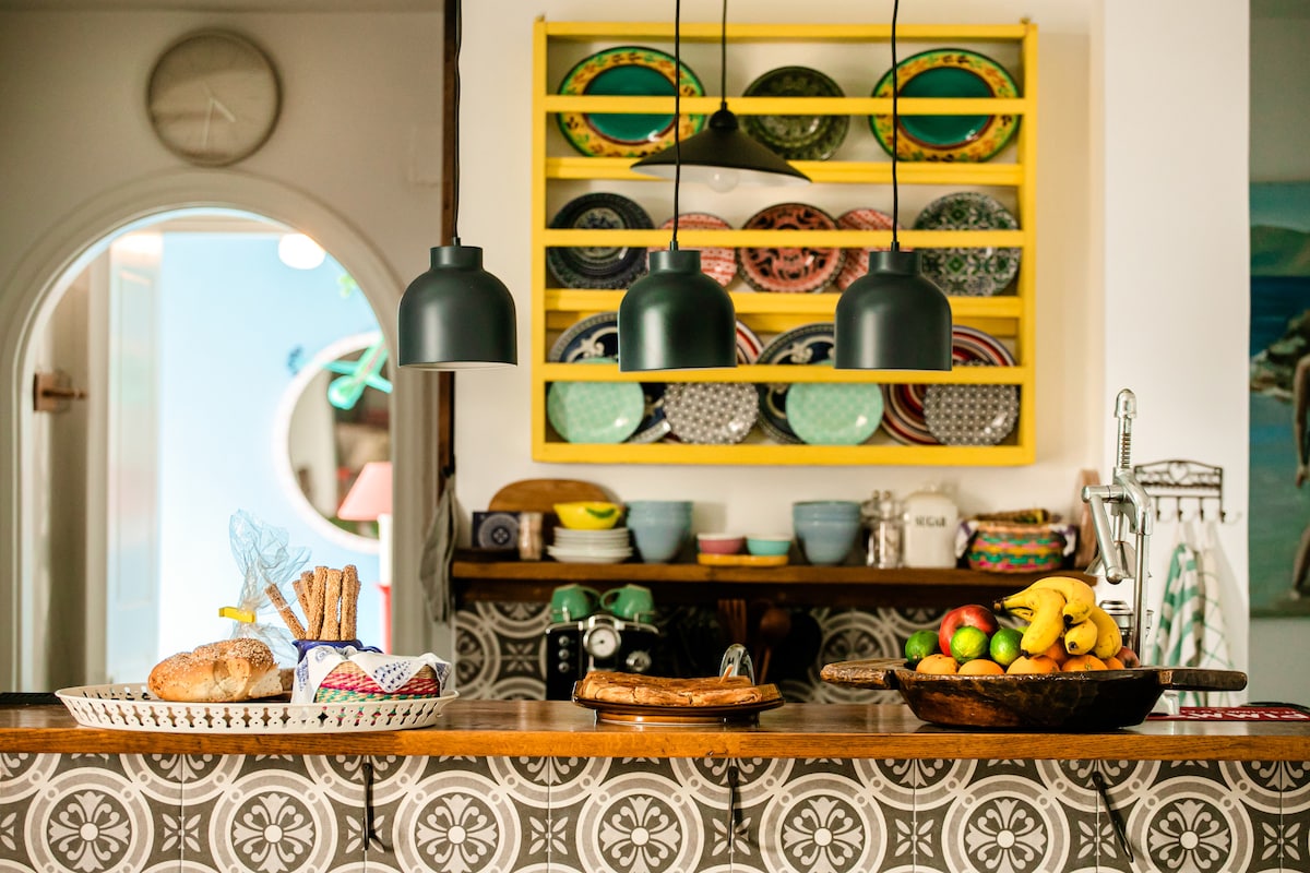 A well-equipped kitchen features a wooden countertop adorned with various fruits, pastries, and decorative items. Above, a vibrant yellow shelf displays an array of colorful dinnerware. Three pendant lights hang above the counter, creating a functional yet inviting cooking space.