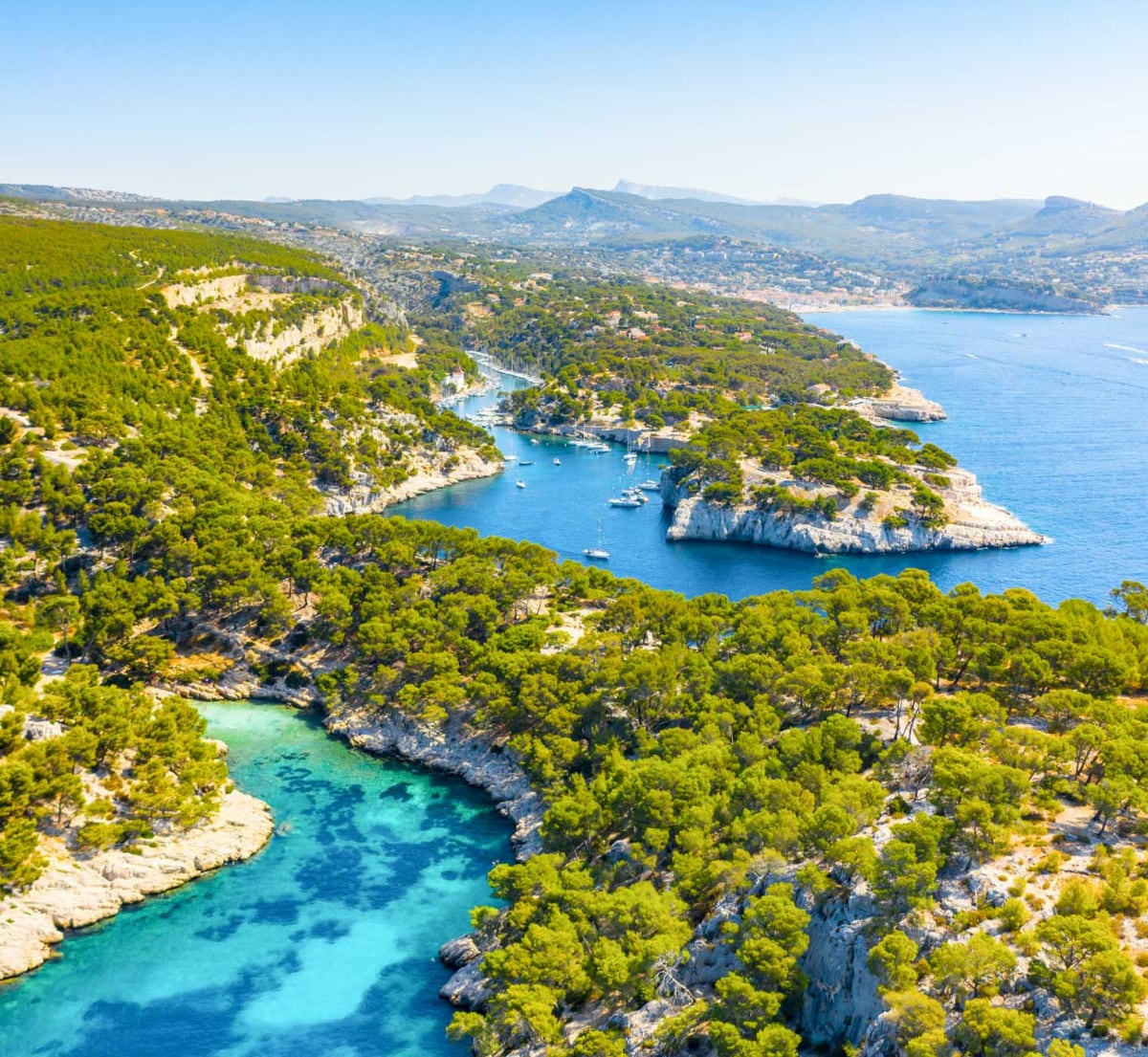 A panoramic view of a coastal landscape, featuring lush green hills interspersed with natural waterways and rocky inlets. Multiple boats are visible navigating the turquoise waters, surrounded by a serene atmosphere. The distant hills provide a backdrop against a clear blue sky.