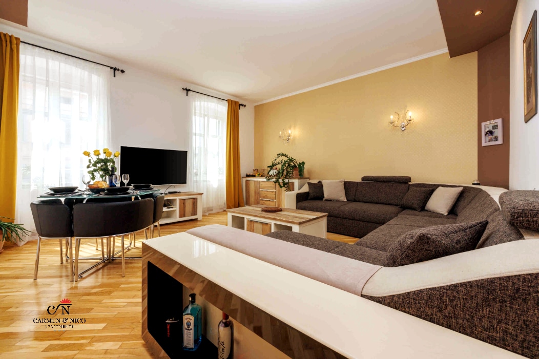 A spacious living room features a large sectional sofa facing a flat-screen television. Natural light pours in through tall windows adorned with light curtains. A dining table with chairs is positioned nearby, accompanied by decorative plants and contrasting wall colors.