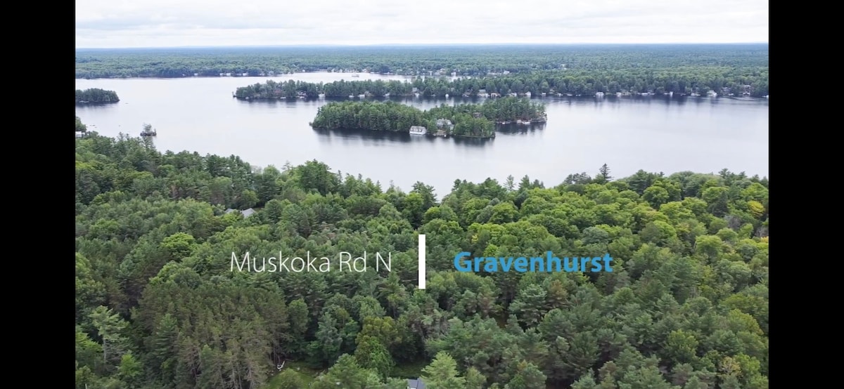 An aerial view showcases a lush green landscape surrounding a tranquil lake. The tree canopy offers varying shades of green, while the body of water reflects the overcast sky. The Gravenhurst area is indicated by a sign, blending seamlessly into the natural scenery.