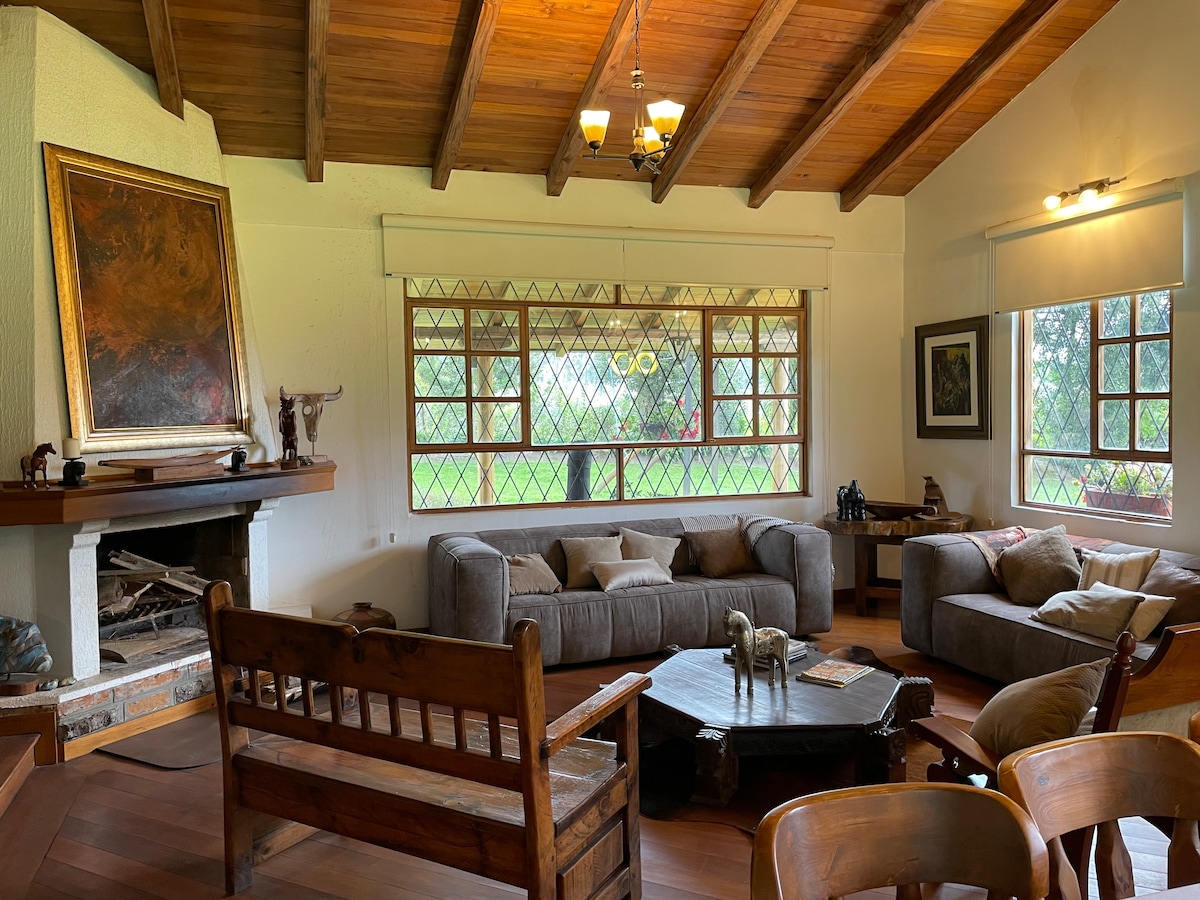 A spacious living area is highlighted by large windows, allowing natural light to fill the room. Comfortable sofas are arranged around a wooden coffee table, and a fireplace provides a focal point alongside decorative art. Warm wooden beams accent the ceiling.