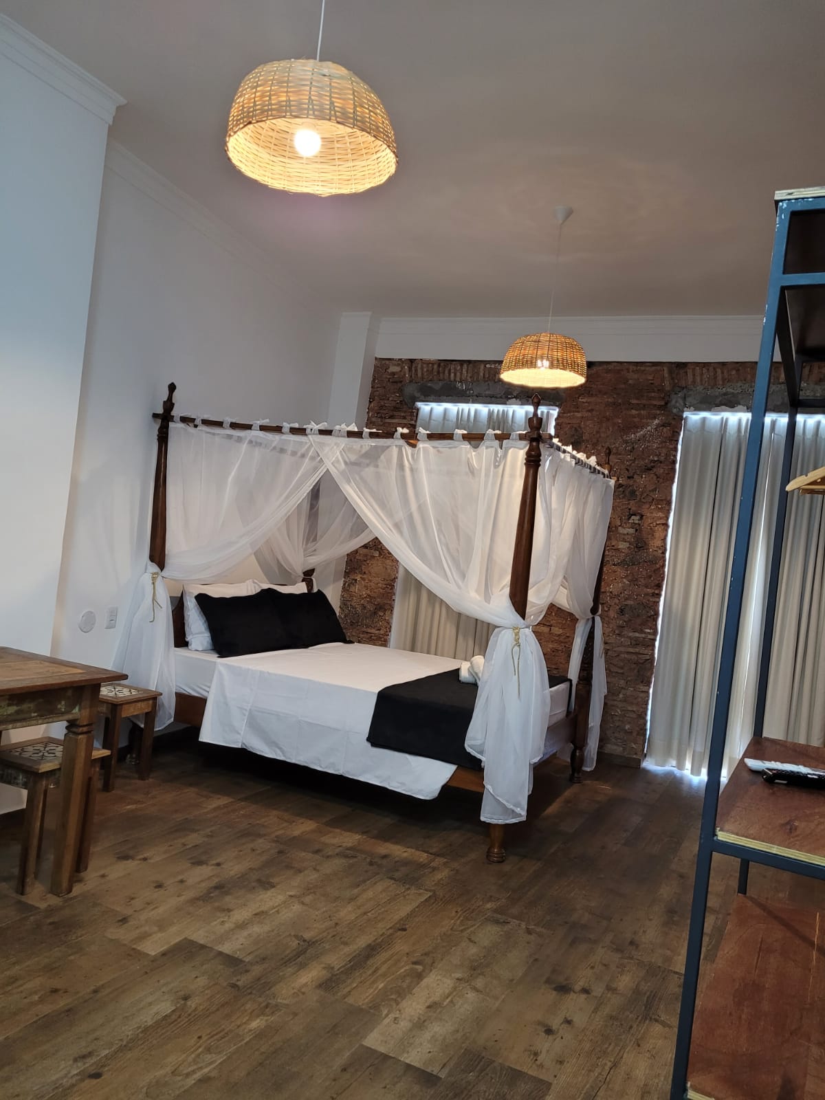 A spacious room is furnished with a wooden canopy bed dressed in white and black linens. Natural light filters through sheer curtains, illuminating the rustic stone accent wall. A dining table made of wood complements the overall decor, enhanced by woven pendant lighting above.