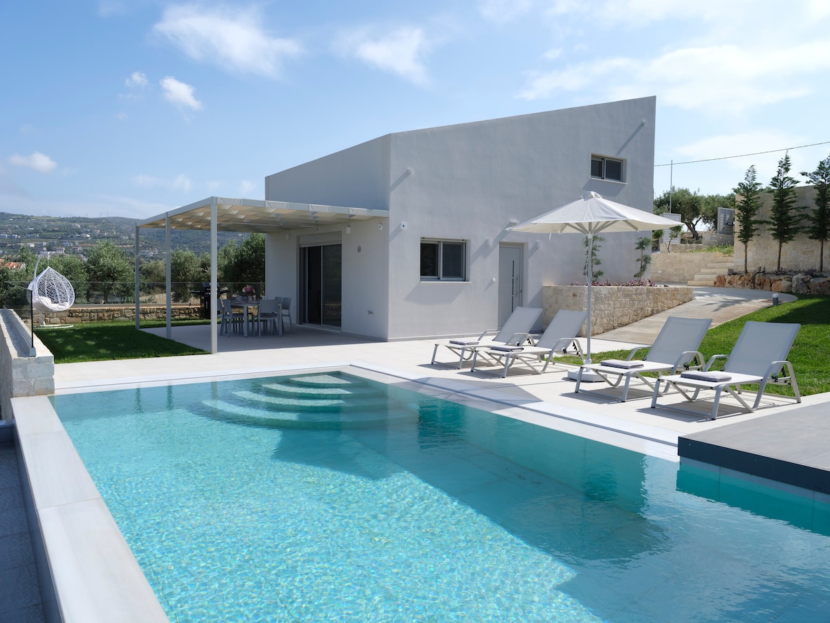 A modern villa is set against a backdrop of green hills and blue skies. The private swimming pool reflects the sunlight, surrounded by comfortable sunbeds and umbrellas. An outdoor dining area is visible under a shaded pergola, enhancing the appeal of the spacious garden.
