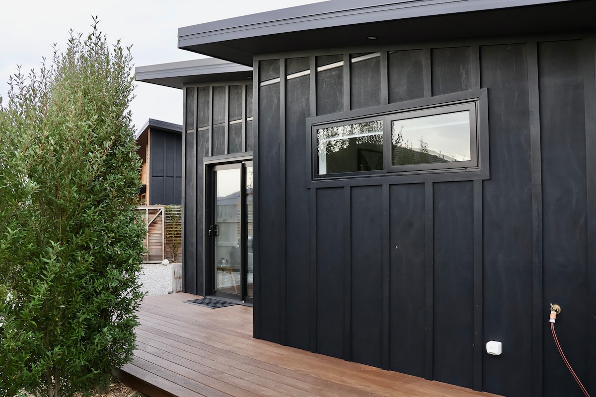 The exterior of the apartment is depicted, showcasing a modern black façade with large windows. A wooden deck extends in front, allowing easy access. Lush greenery surrounds the entrance, adding a touch of nature to the contemporary design.