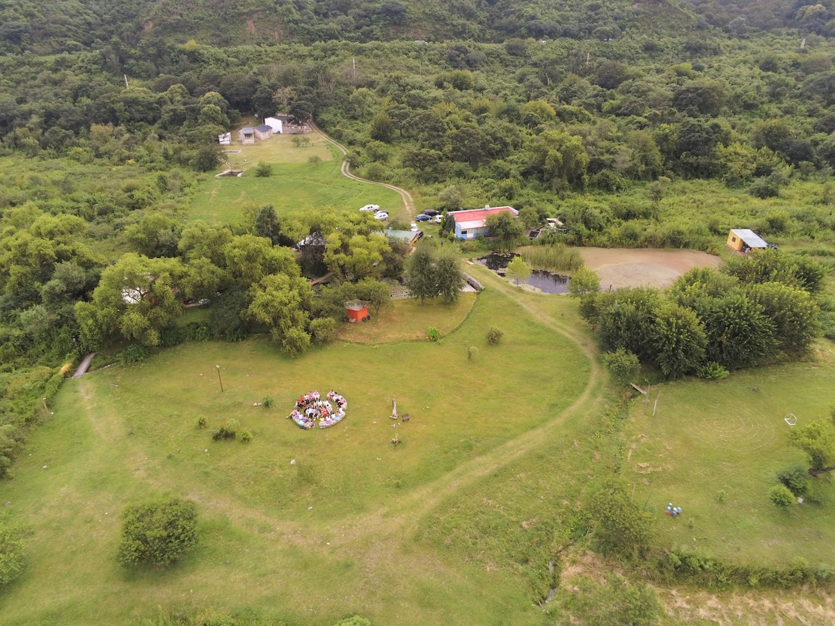 An aerial view captures a vast green landscape, featuring a circular gathering area surrounded by trees. Various pathways meander through the grass, leading towards distant structures and a small pond, all set within a tranquil and expansive natural environment.