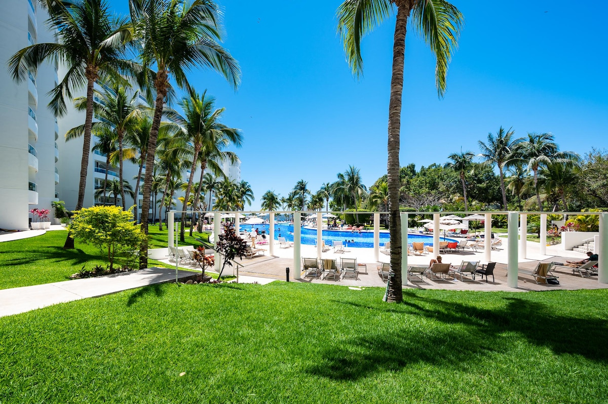 The image showcases a vibrant outdoor area featuring well-maintained green lawns and palm trees. A sparkling pool is visible in the background, surrounded by lounge chairs and shaded umbrellas, providing a refreshing oasis for relaxation.