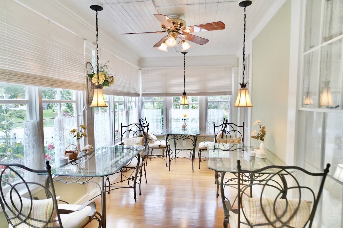 A bright sunroom features a light wooden floor and white-paneled ceiling. Glass-top tables are surrounded by wrought iron chairs, with soft cushions providing comfort. Natural light floods through large windows adorned with curtains, enhancing the room's airy ambiance.