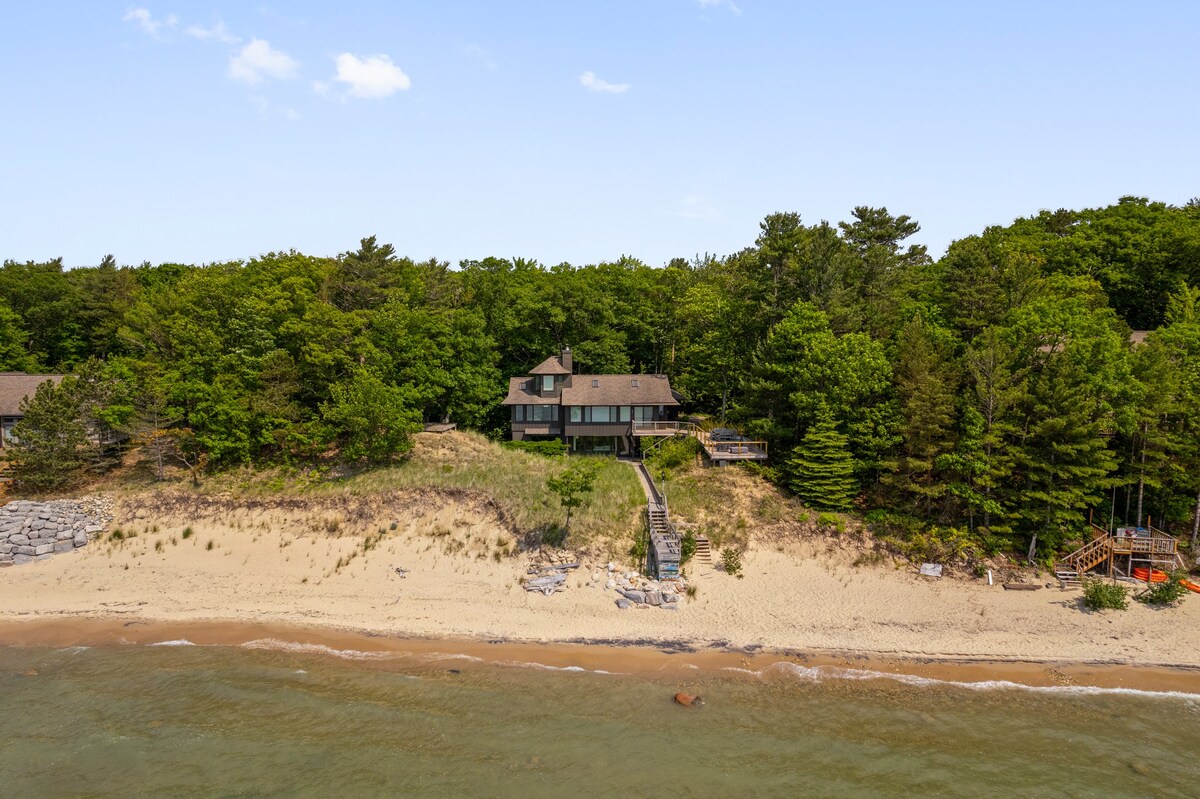The property is nestled along the sandy shore of East Bay, Lake Michigan, surrounded by lush trees. A staircase leads from the home down to the beach, offering easy access to the water. The building features a well-integrated design with natural surroundings.