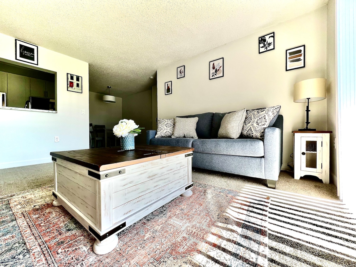 A spacious living area features a gray sofa adorned with patterned pillows, complemented by a coffee table with a rustic finish. Natural light fills the room through large windows, casting soft shadows across a decorative area rug.