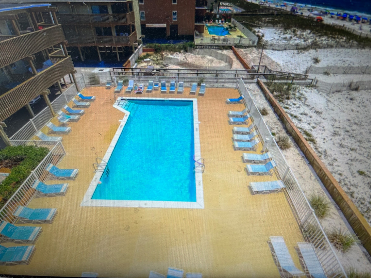 An aerial view of the pool area showcases a spacious swimming pool surrounded by a deck lined with lounge chairs and umbrellas. The area is framed by sandy beach visible in the background, along with buildings of the resort on either side.