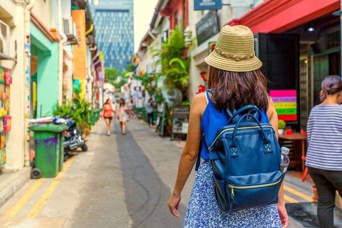 A person is seen walking down a vibrant street lined with colorful shop houses. The scene features a mix of greenery and urban elements, with distant modern buildings visible, suggesting a lively blend of tradition and contemporary life.