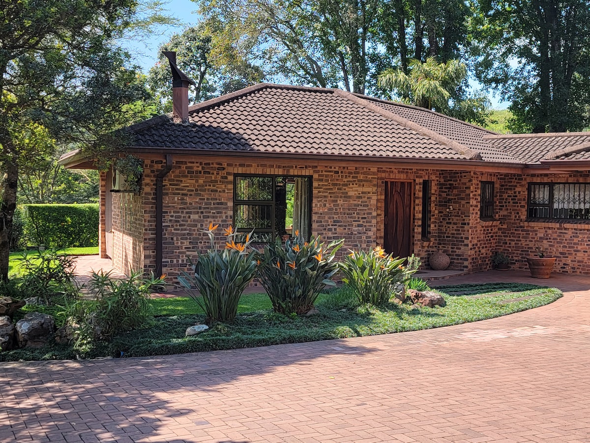 The exterior of the cottage features a brick façade surrounded by lush greenery and tropical plants. A neatly paved pathway leads to the entrance, showcasing a tranquil atmosphere. Tall trees provide shade, contributing to the serene outdoor environment.