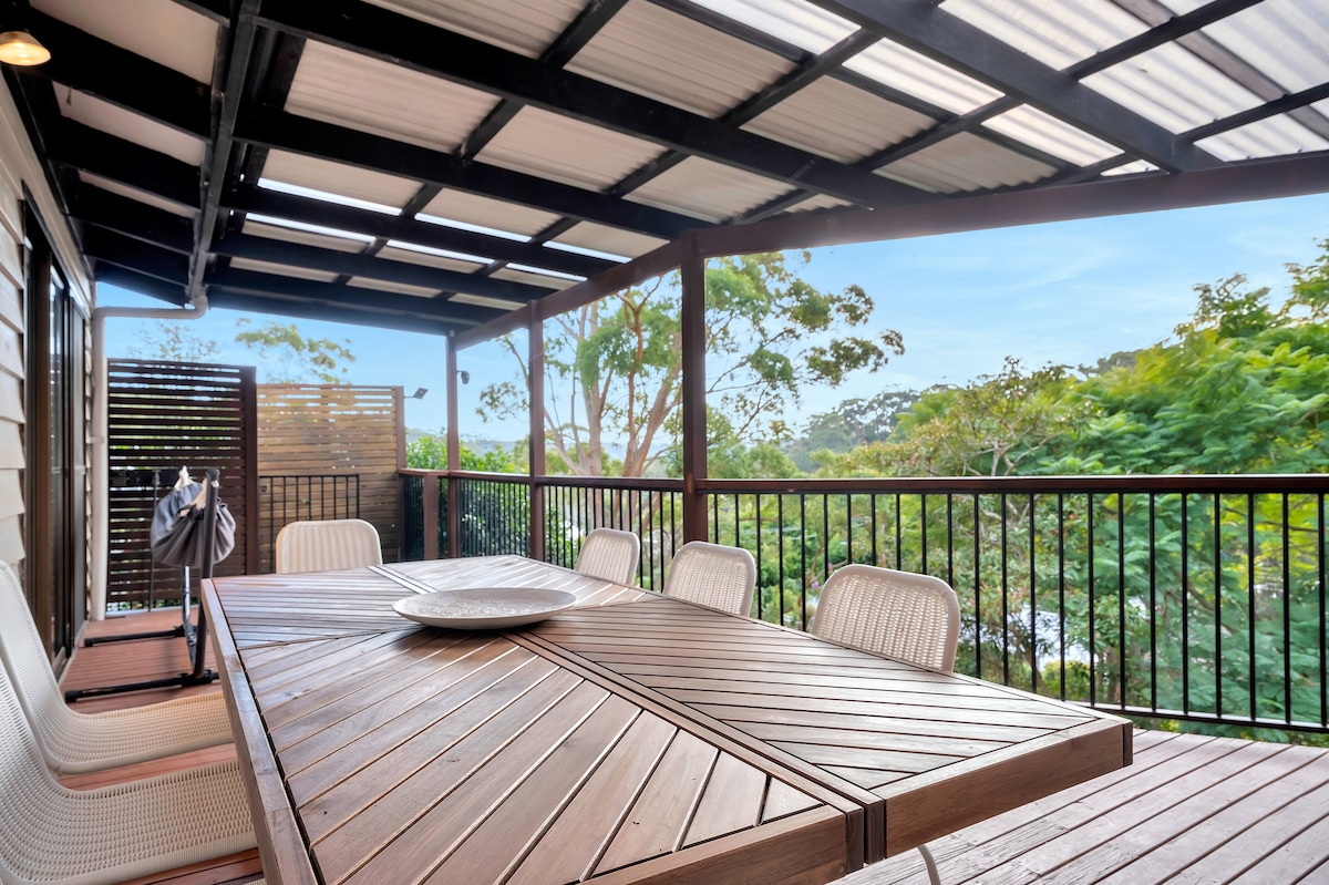 An outdoor deck is featured with a large wooden dining table surrounded by several white chairs. Lush greenery is visible beyond the railing, providing a serene backdrop. Shade is offered by a structured overhang, enhancing the space for al fresco dining.