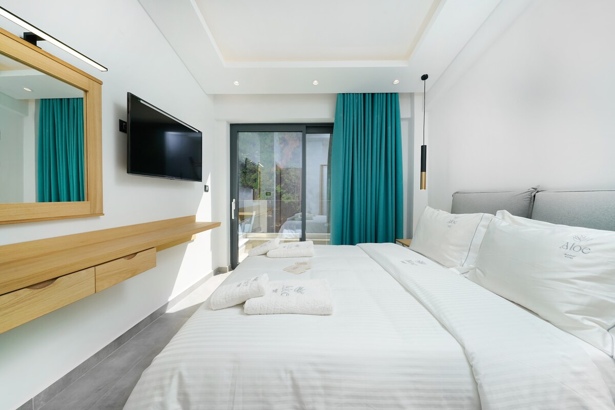 A bright and spacious bedroom features a king-size bed with crisp white linens. Large glass doors provide access to an outdoor area, while turquoise curtains frame the windows. A wall-mounted television and a wooden dresser add functionality to the room.