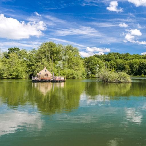 A floating cabin is depicted on calm waters, surrounded by lush greenery. The structure features a natural wood design, harmonizing with the landscape. Bright blue skies with scattered clouds reflect on the water, creating a serene atmosphere in this secluded environment.