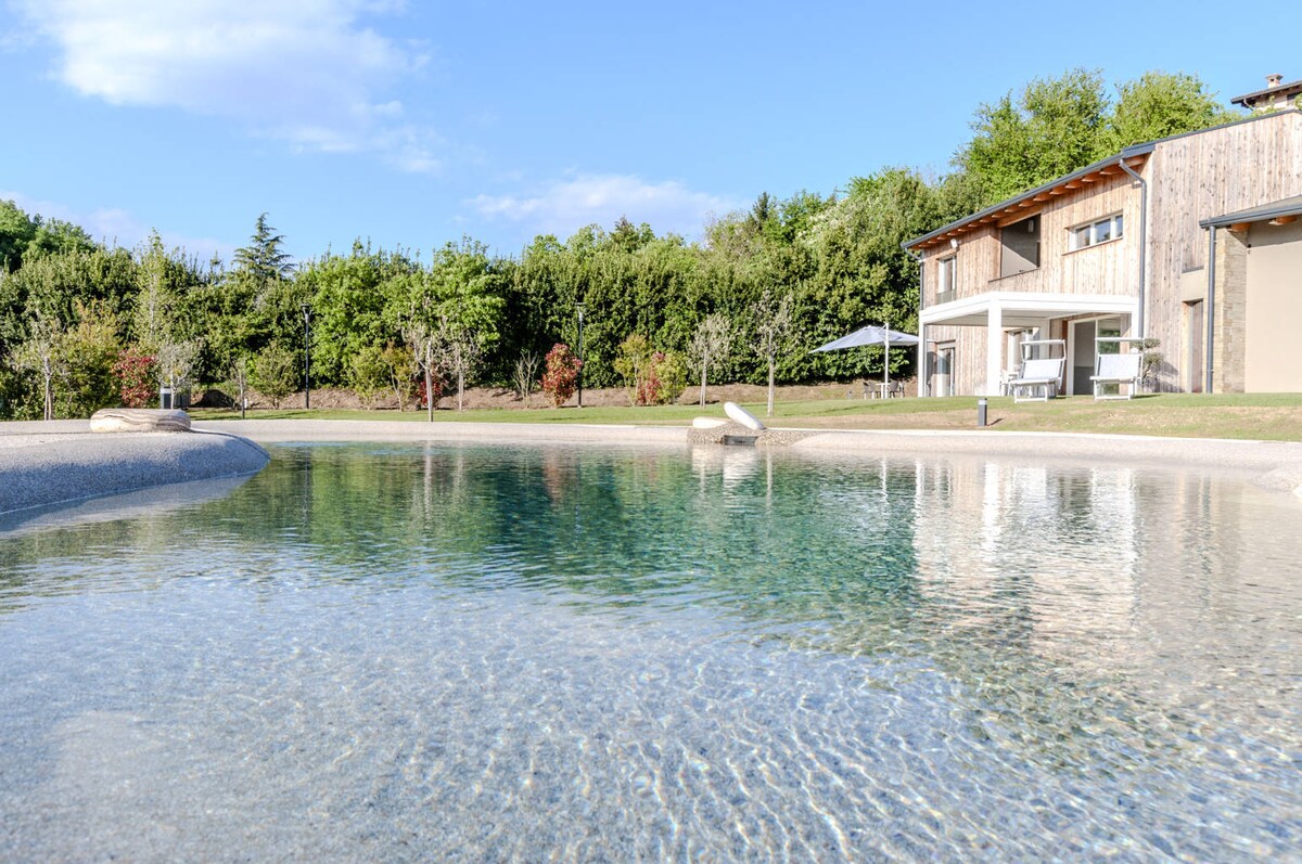 An inviting outdoor pool is surrounded by a landscaped area featuring trees and green grass. The pool displays a gentle gradient from shallow to deeper sections, with smooth stones incorporated into the design. The backdrop includes a modern building with outdoor seating under a shaded area.