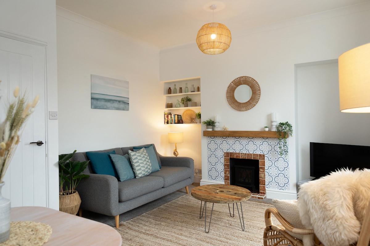 A modern living area is featured, showcasing a grey sofa with decorative cushions and a wood coffee table. A statement light fixture hangs overhead, and a bright wall contains shelves displaying various decorative items. A woven rug anchors the space, complemented by a stylish fireplace adorned with tiles.