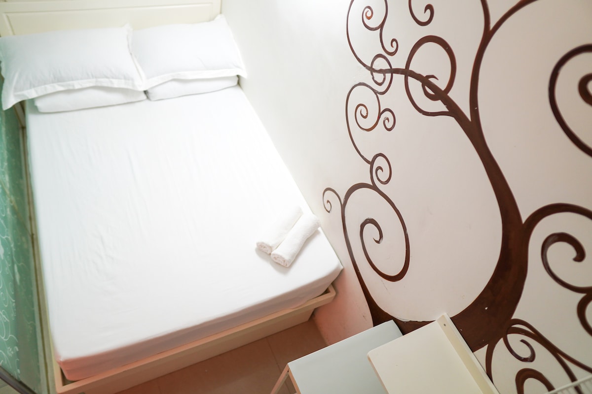 A sleeping area features a spacious bed with clean white linens and two pillows placed at the head. A small stack of towels rests on the edge. A decorative tree design adorns the wall, adding a touch of artistry to the room.
