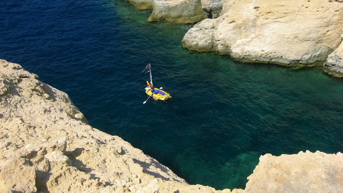 A small yellow raft floats in calm blue waters, surrounded by rugged rocky cliffs. The clarity of the water reflects varying shades of blue and green, while the sun illuminates the stark, sandy tones of the cliffs nearby.