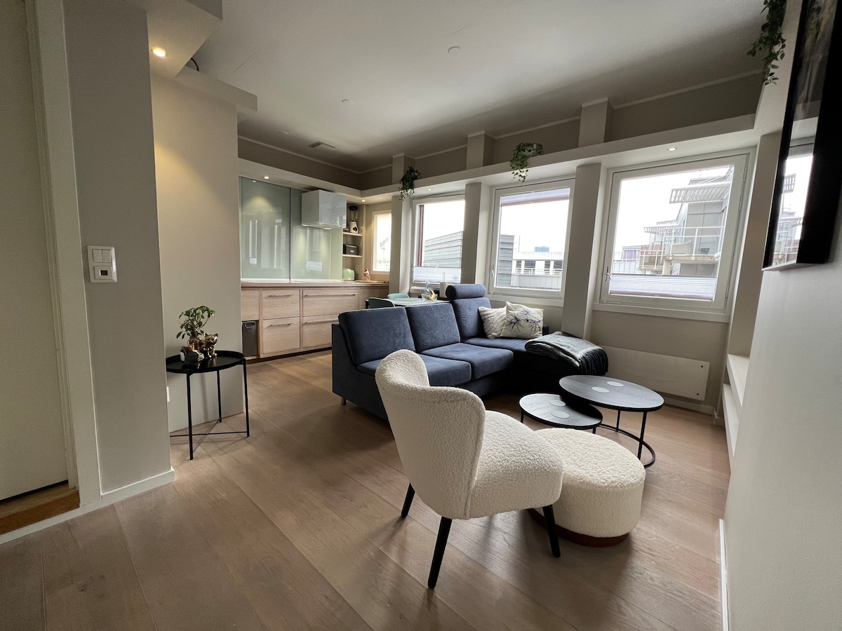 A comfortable living area features a spacious sofa in a soft blue hue, complemented by a light armchair. A round coffee table sits in front of the sofa, while natural light enters through large windows, illuminating the modern design of the space.