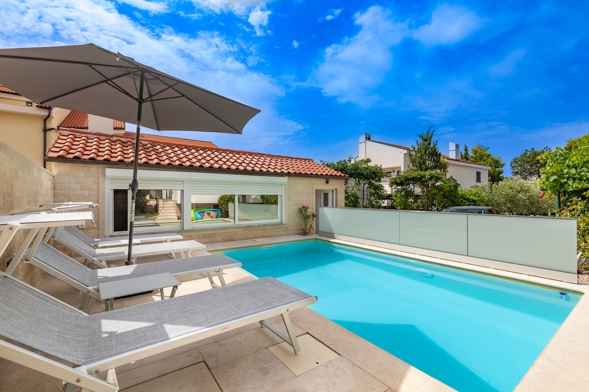 A shared pool area features a clear blue pool with sun loungers positioned around it. An umbrella provides shade over one of the loungers. Lush greenery is visible in the surrounding area, with a glimpse of the house reflected in the pool's calm surface.