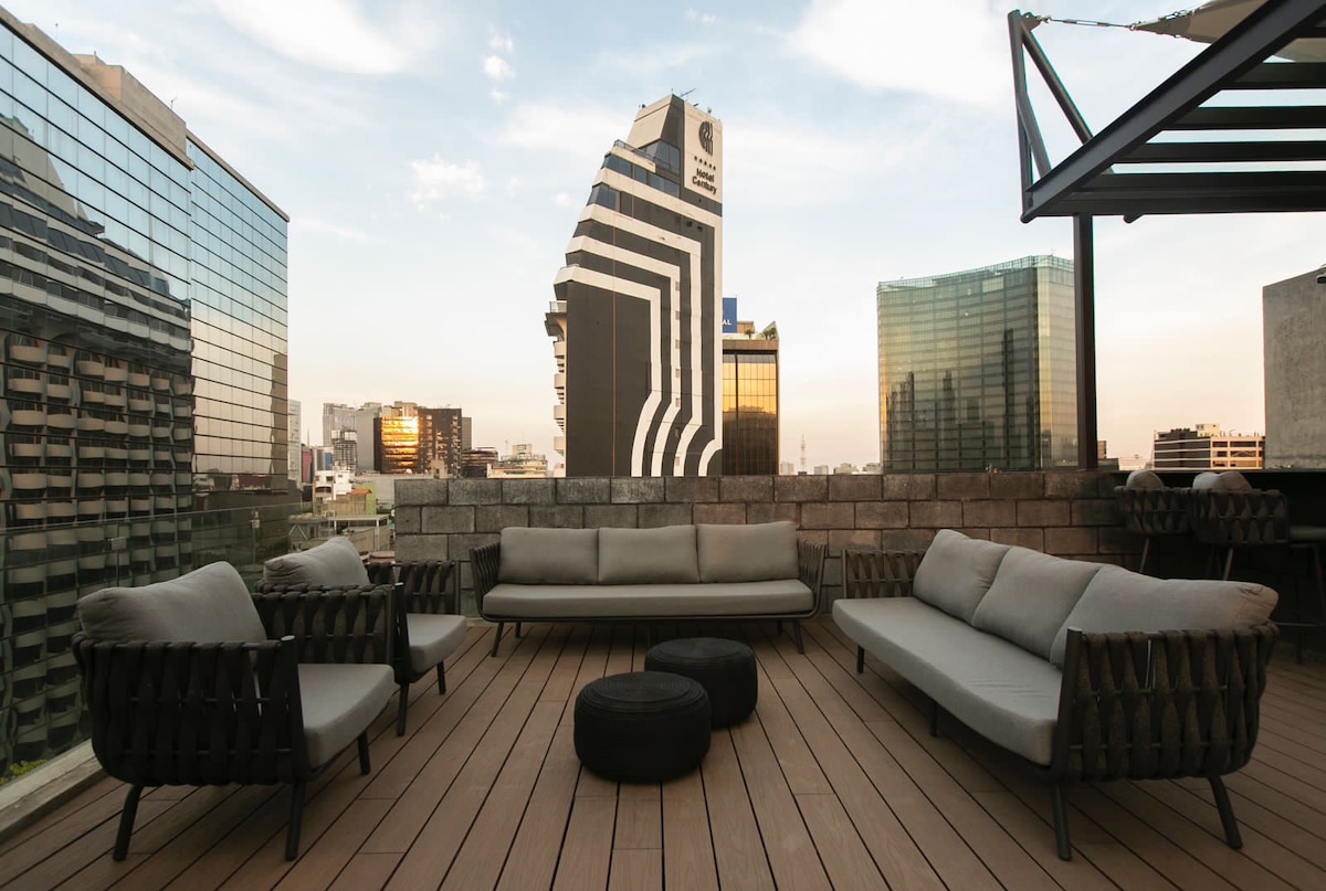 A spacious rooftop terrace features comfortable seating arrangements, including a large sofa and two chairs with cushioned backs. A wooden deck provides a warm backdrop. Towering glass buildings outline the skyline, reflecting the soft glow of sunset.