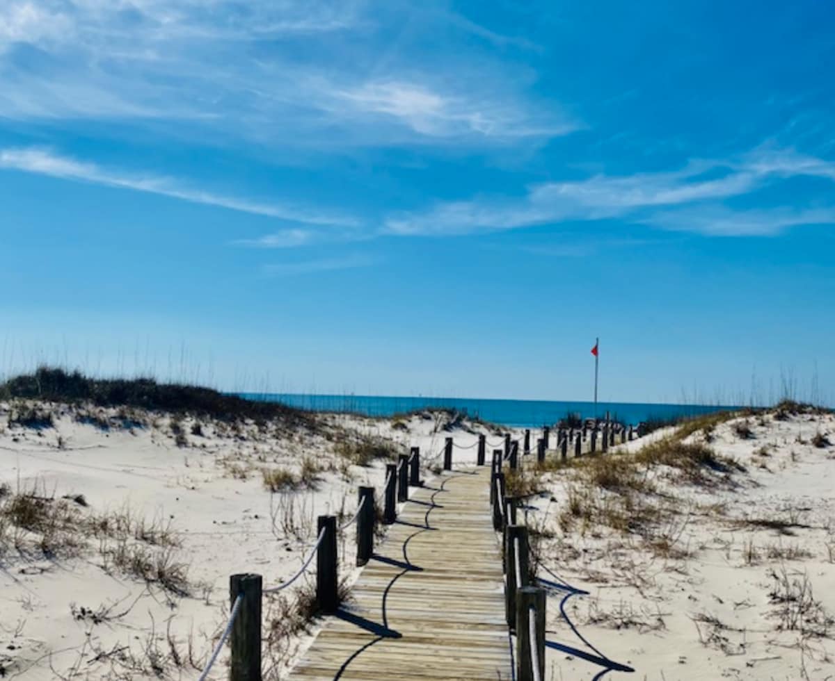 A wooden boardwalk leads through sandy terrain towards the ocean. Dune grass flanks the path, and a flag marks the beach entrance. The sky is clear, with gentle clouds present, creating a serene backdrop for the coastal view.