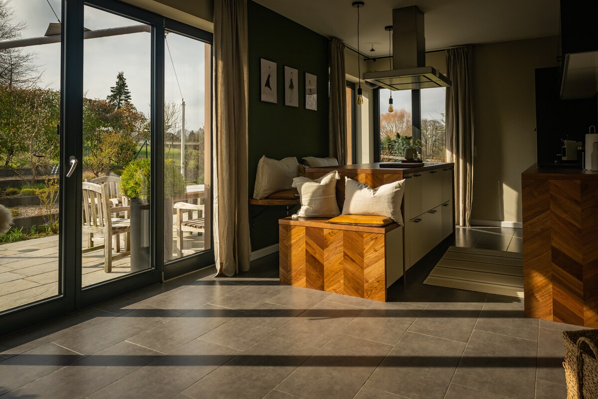 A spacious kitchen area features large glass doors that open to the outdoor terrace. A wooden island with seating provides a gathering space, while natural light floods the room through the windows. Neutral tones and modern design elements create a welcoming atmosphere.