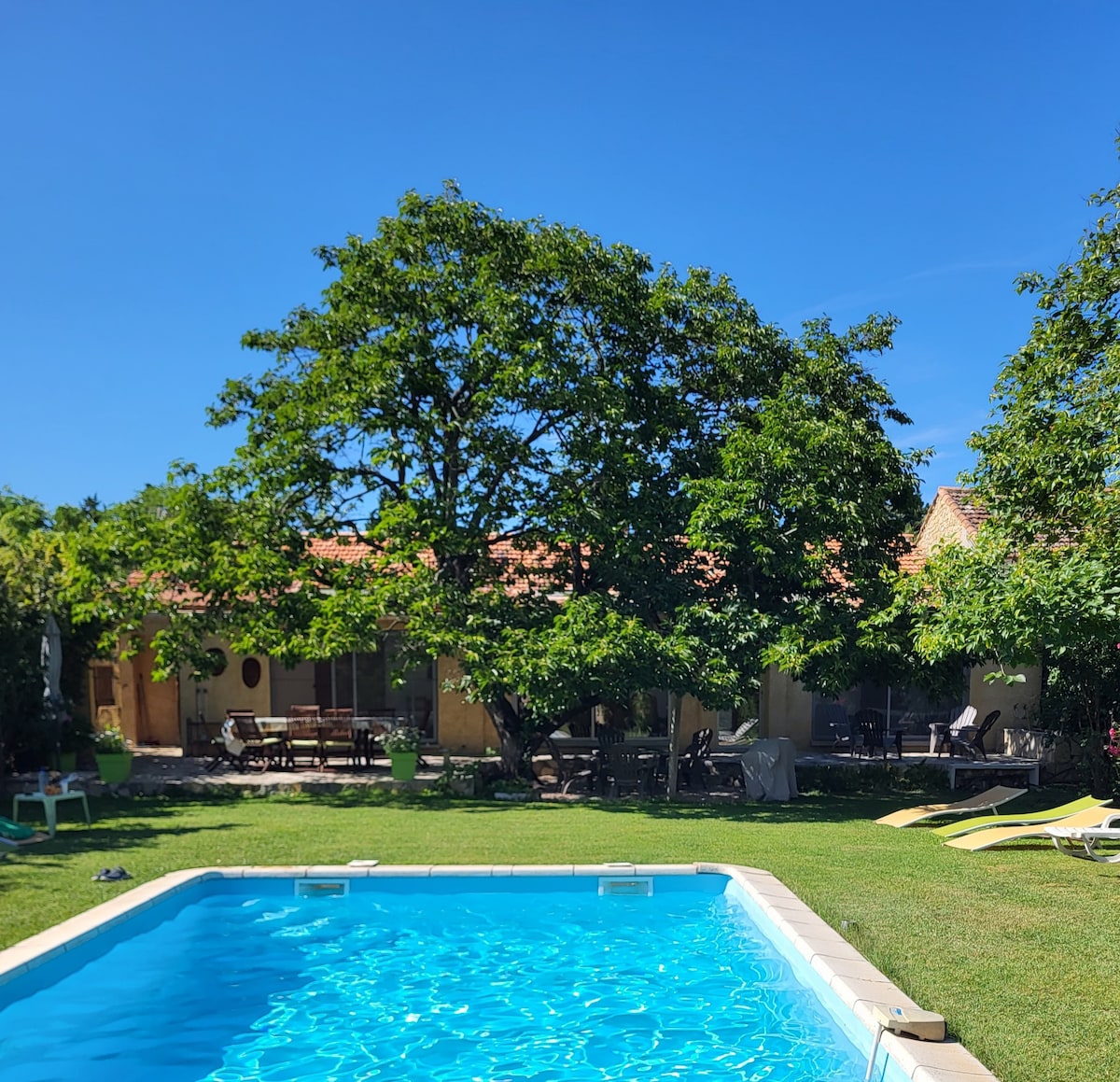 A clear view of a sparkling blue pool framed by lush greenery and large trees. The villa is visible in the background, showcasing a spacious terrace with seating arrangements. The well-maintained garden offers a tranquil setting for relaxation.