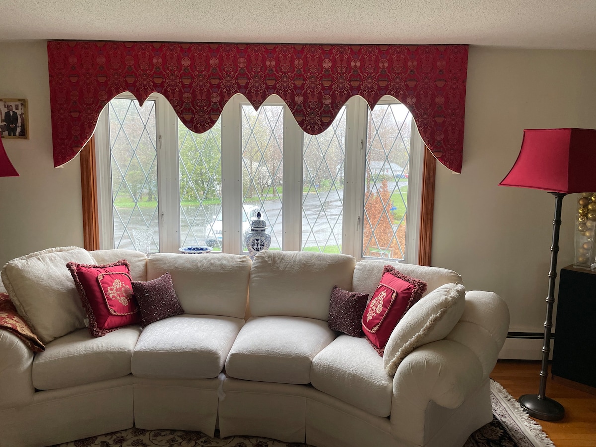 A spacious white sectional sofa is positioned near a large bay window. The window is adorned with a decorative valance and has soft red curtains on the sides. Two red lamps with white shades provide warm lighting, complementing the cozy ambiance.