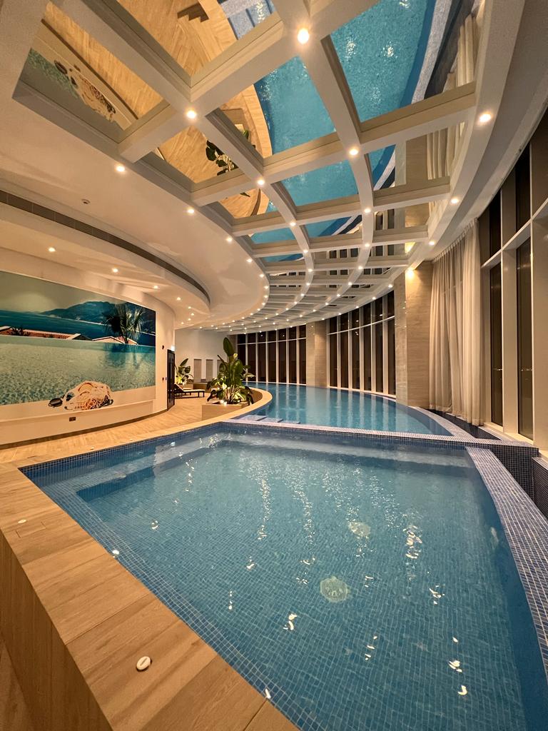 An indoor infinity pool is featured with a curved edge, surrounded by large glass windows allowing natural light to fill the space. Decorative plants and elegant seating areas enhance the tranquil setting. A mural depicting a serene ocean scene is displayed on the wall.