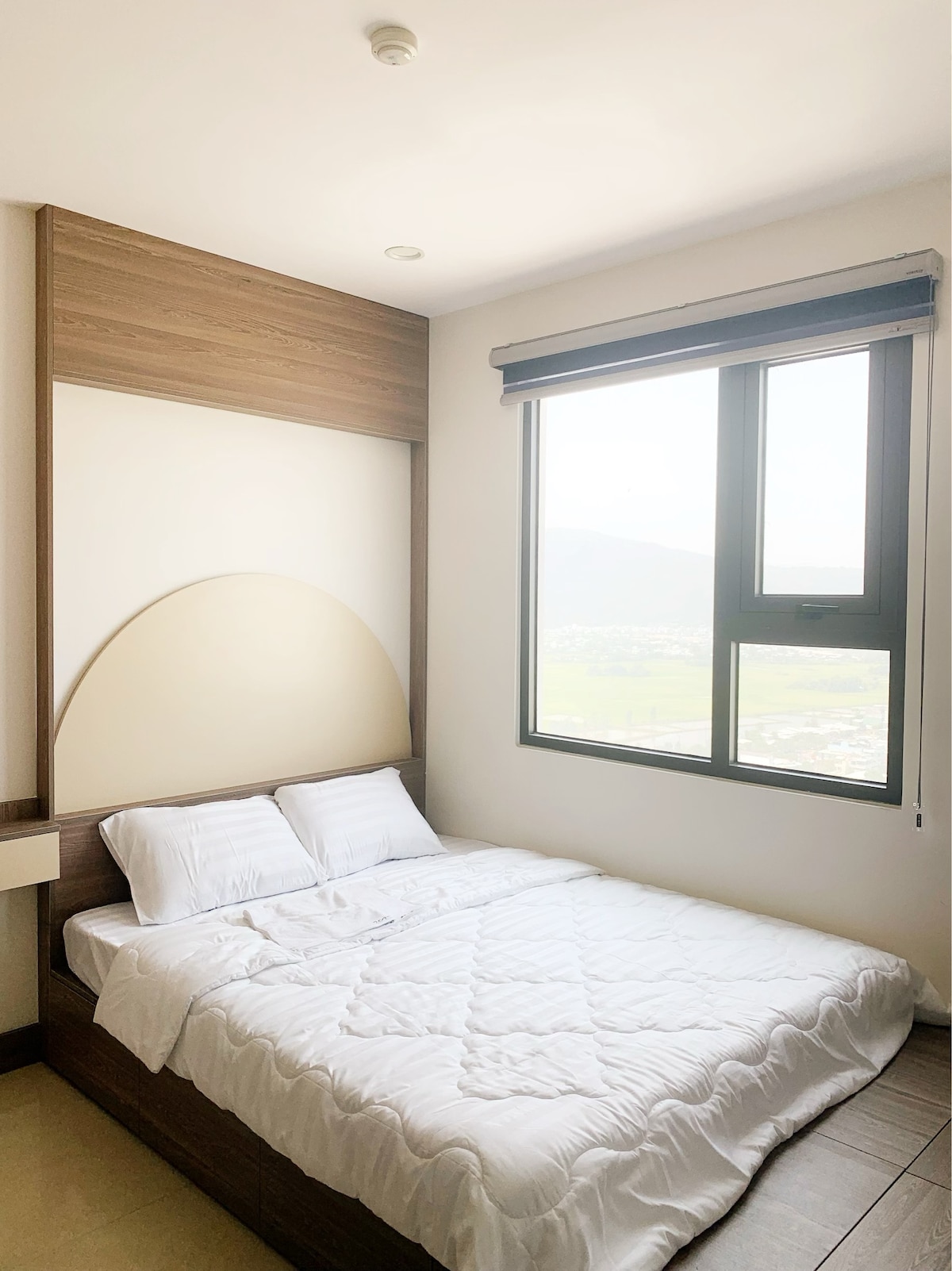 A bright bedroom features a spacious bed with a white quilt. Large windows allow natural light to fill the room, showcasing scenic views of the surrounding landscape. A wooden accent wall adds warmth to the minimalist decor.