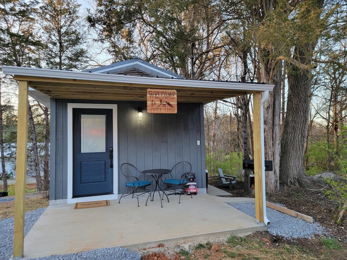 A charming tiny home is displayed with a covered porch featuring a small table and two chairs. The front door is painted blue, and a wooden sign hangs above. Surrounding greenery and trees enhance the natural setting.