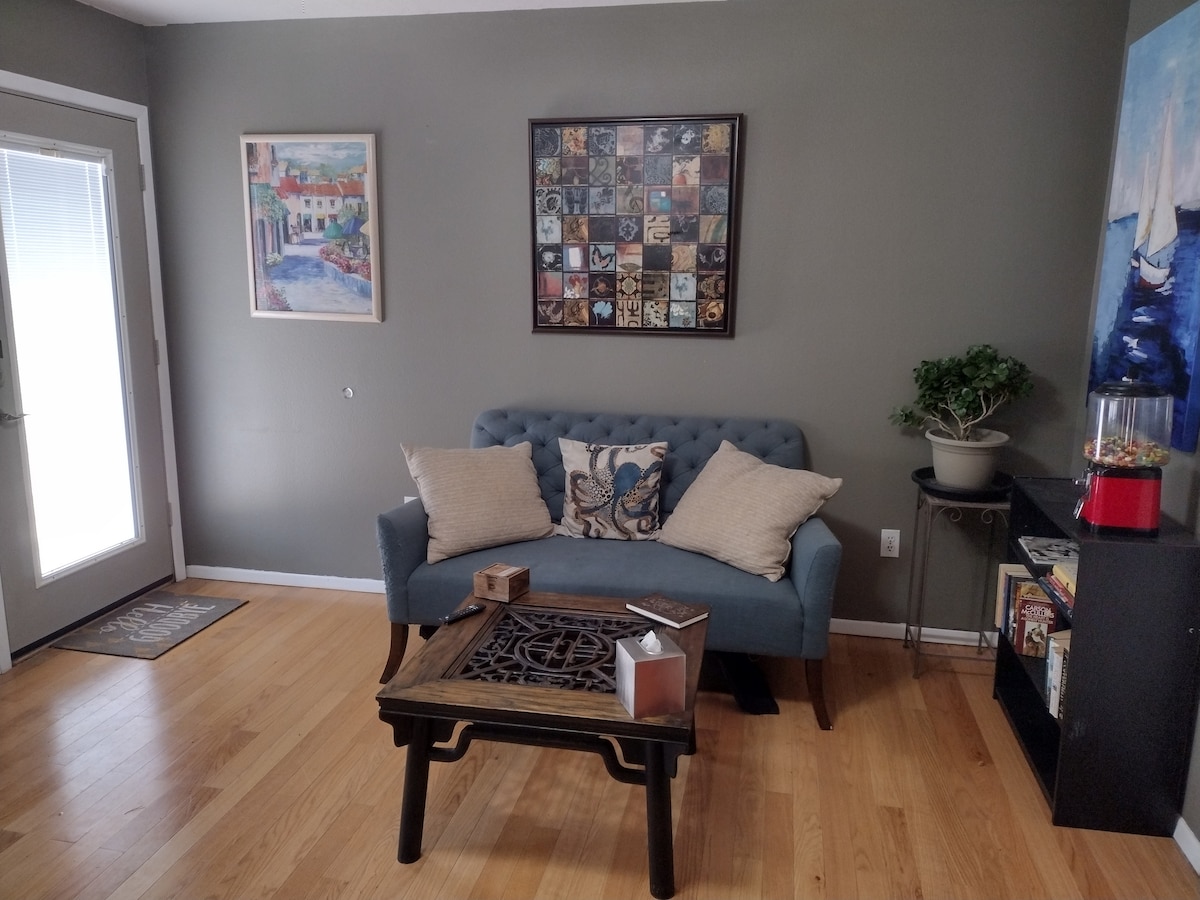 A well-furnished front room features a tufted blue couch with decorative pillows. A wooden coffee table with intricate detailing occupies the center. Artwork hangs on the walls, while a small plant adds a touch of greenery. Light enters through a glass door leading outside.